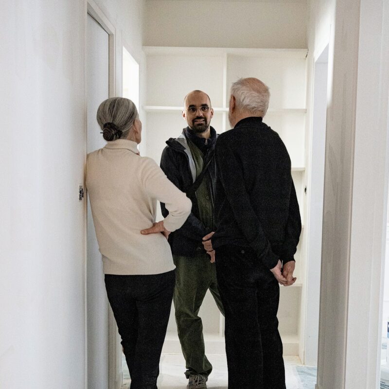 Victor discute avec un couple de personnes âgées dans un couloir aux murs blancs d'un logement en travaux